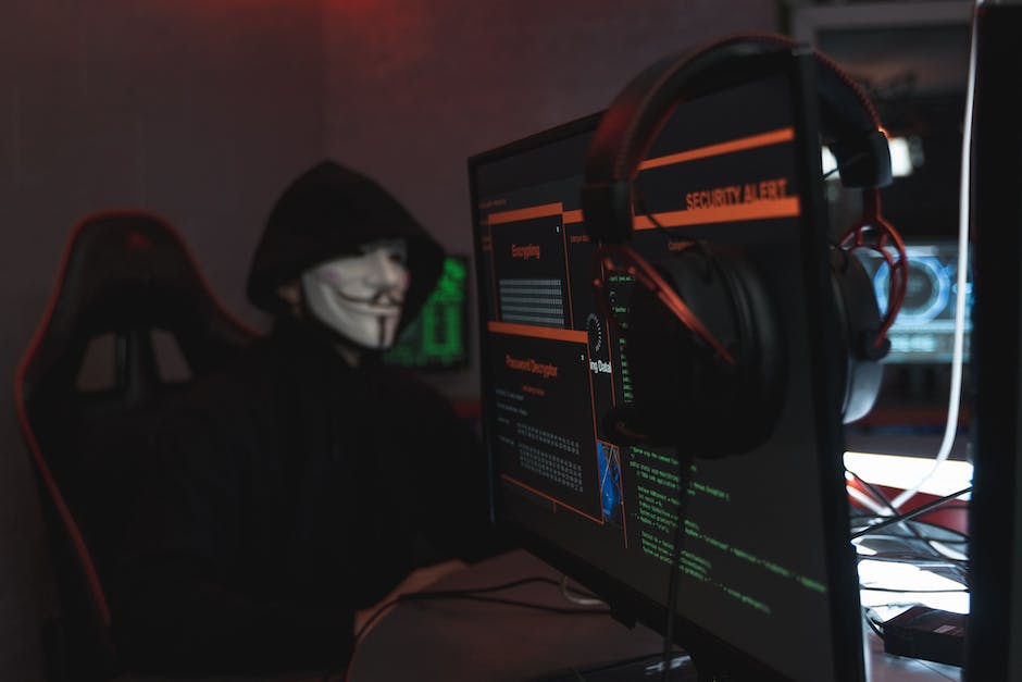 Image depicting a person wearing a black hoodie sitting in front of multiple computer screens with lines of code, illustrating cybercrime activities.